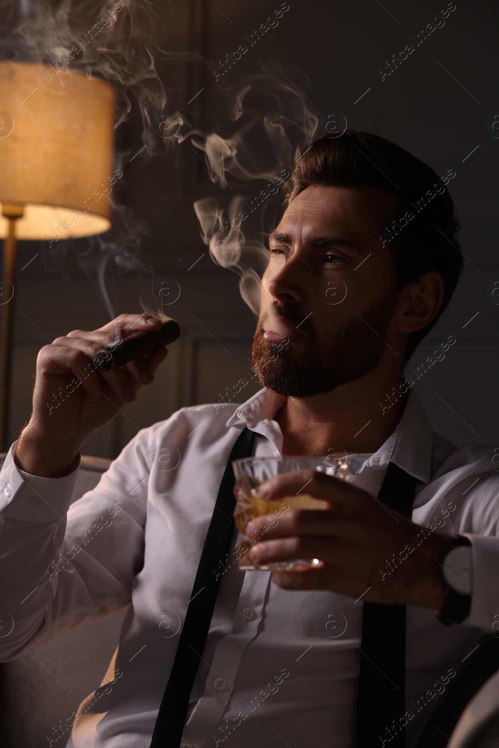 Photo of Handsome man with glass of whiskey smoking cigar at home