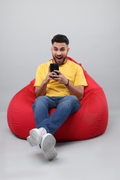 Photo of Emotional young man using smartphone on bean bag chair against grey background