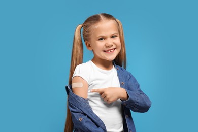 Photo of Happy girl pointing at sticking plaster after vaccination on her arm against light blue background