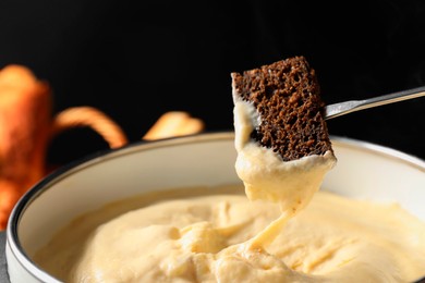 Photo of Dipping piece of bread into fondue pot with melted cheese on black background, closeup