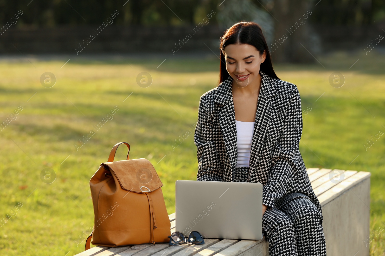 Photo of Beautiful young woman with stylish backpack and laptop on bench in park, space for text