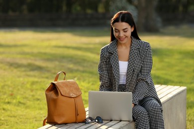 Beautiful young woman with stylish backpack and laptop on bench in park, space for text