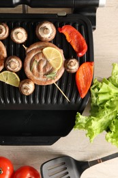 Photo of Electric grill with homemade sausages, vegetables and spatula on wooden table, flat lay