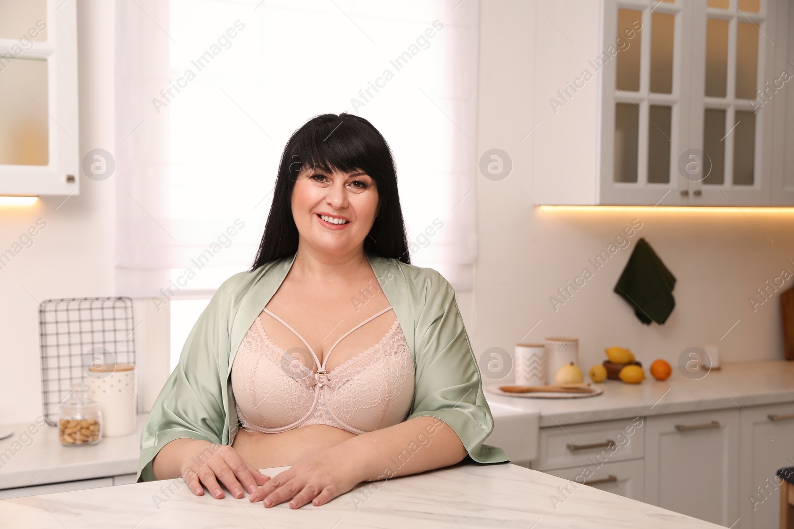 Photo of Beautiful overweight woman in beige underwear and silk robe at kitchen table. Plus-size model