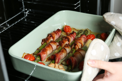 Woman taking ceramic baking dish with bacon wrapped asparagus from oven, closeup