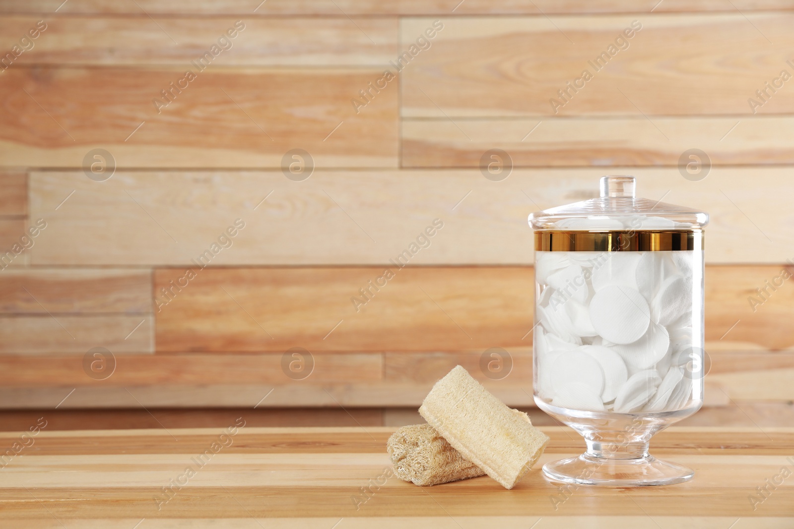 Photo of Decorative glass jar with cotton pads and loofahs on table against wooden background. Space for text