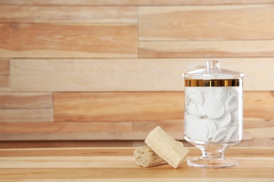 Photo of Decorative glass jar with cotton pads and loofahs on table against wooden background. Space for text