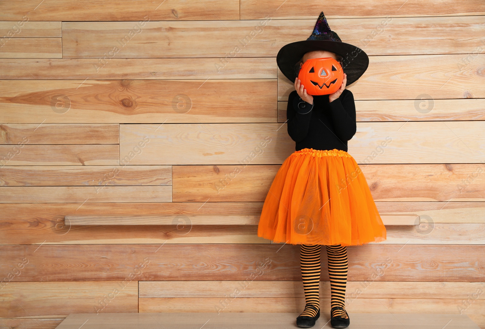 Photo of Cute little girl with pumpkin candy bucket wearing Halloween costume near wooden wall. Space for text