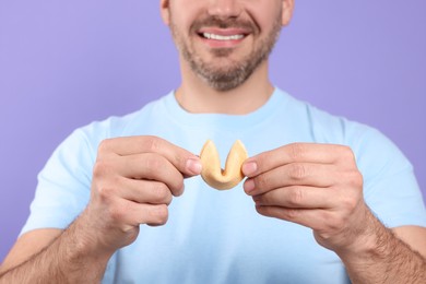Photo of Happy man holding tasty fortune cookie with prediction on violet background, closeup