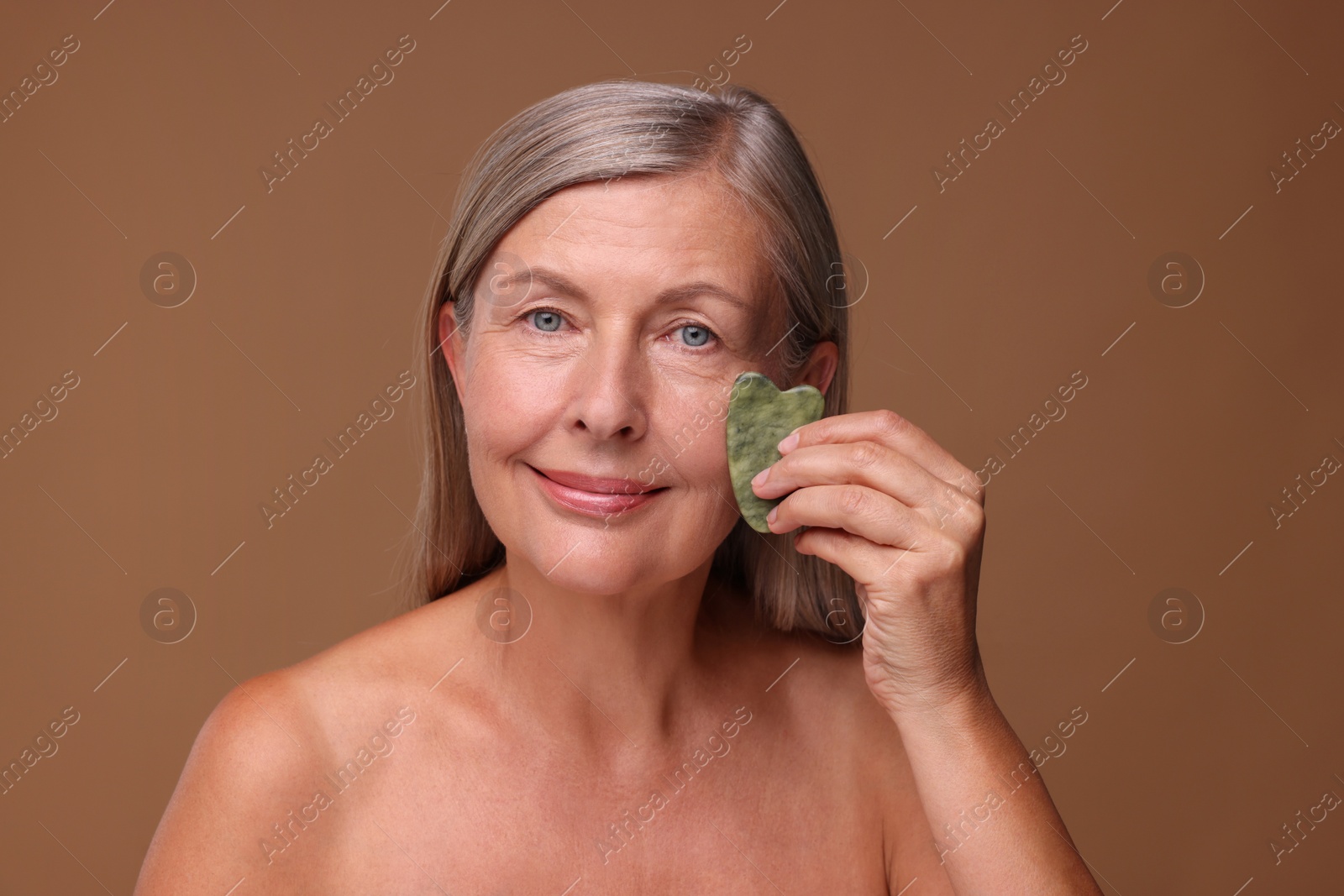 Photo of Woman massaging her face with jade gua sha tool on brown background