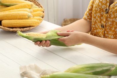 Photo of Woman husking corn cob at white wooden table, closeup