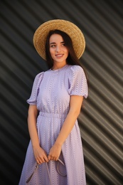 Photo of Beautiful young woman in stylish violet dress and straw hat near dark corrugated metal wall