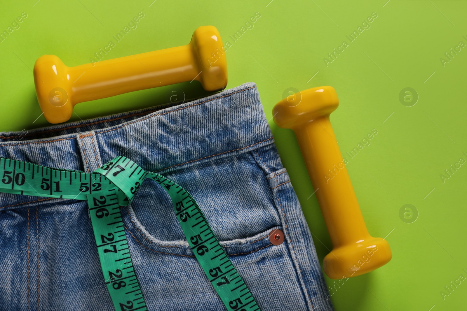 Photo of Jeans, dumbbells and measuring tape on light green background, flat lay. Weight loss concept