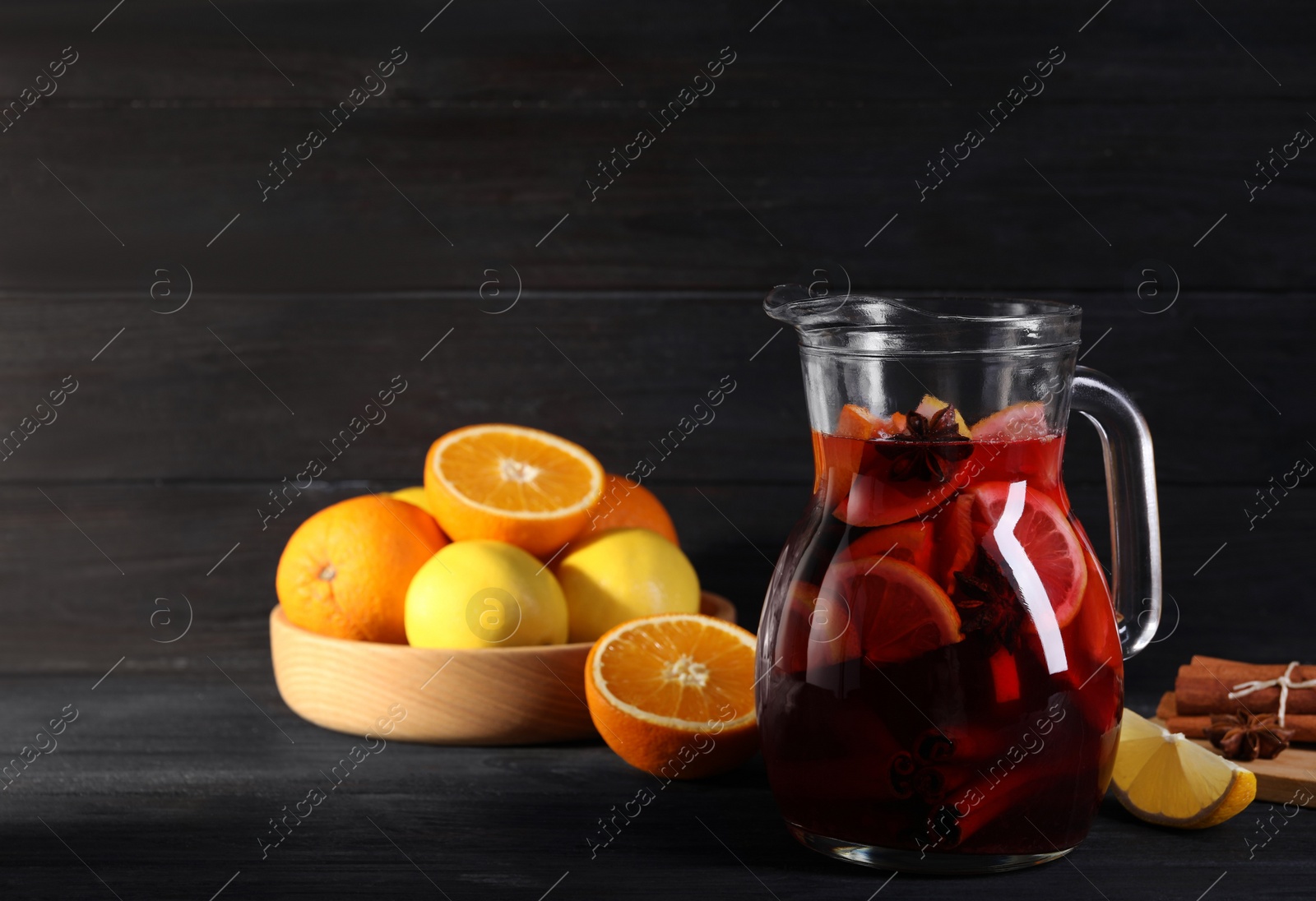 Photo of Glass jug of aromatic punch drink and ingredients on black wooden table. Space for text