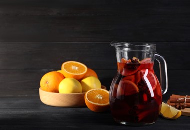 Photo of Glass jug of aromatic punch drink and ingredients on black wooden table. Space for text