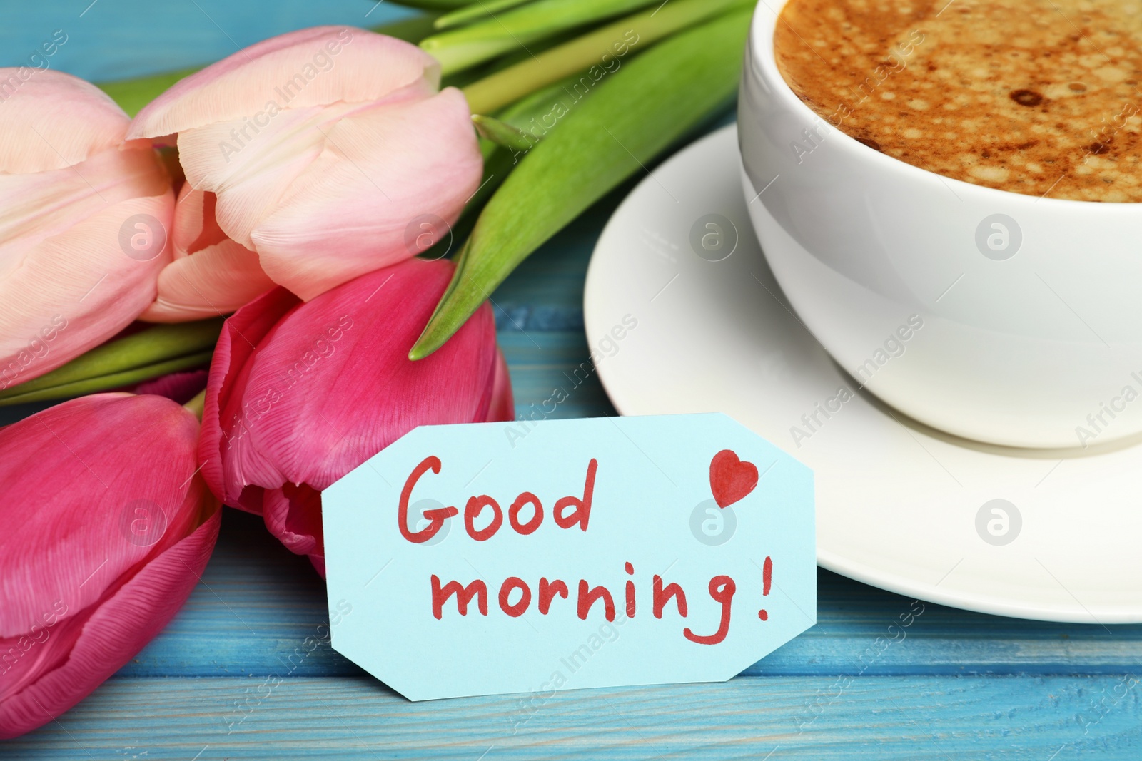 Photo of Cup of aromatic coffee, beautiful pink tulips and Good Morning note on light blue wooden table, closeup