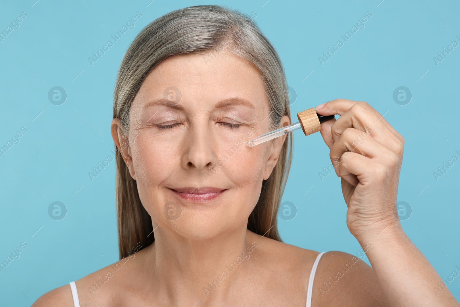 Photo of Senior woman applying cosmetic product on her aging skin against light blue background. Rejuvenation treatment