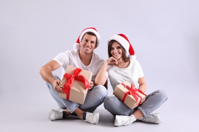 Photo of Beautiful happy couple in Santa hats sitting with Christmas gifts on light background