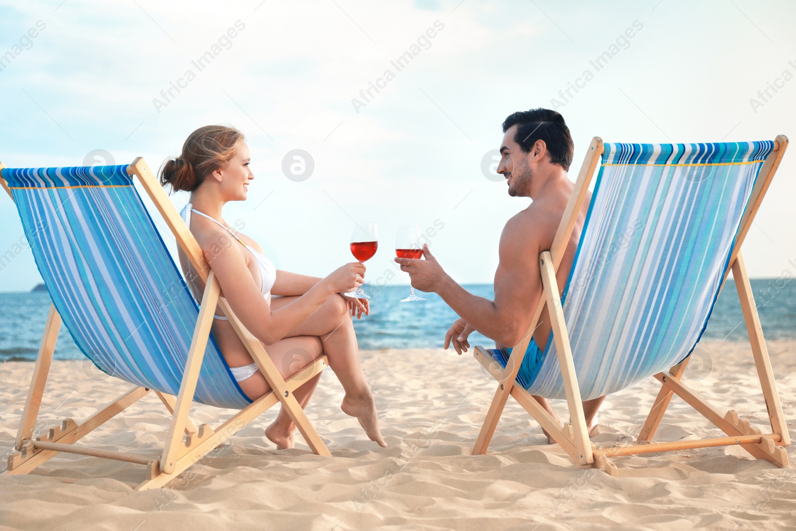 Photo of Happy young couple with glasses of wine sitting on deck chairs at sea beach