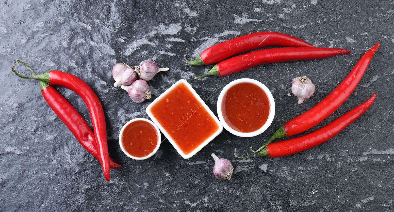 Photo of Spicy chili sauce, peppers and garlic on black textured table, flat lay