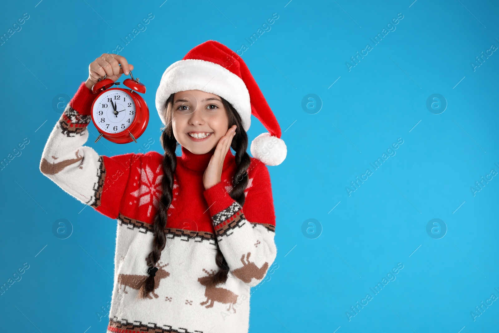 Photo of Girl in Santa hat with alarm clock on light blue background, space for text. New Year countdown