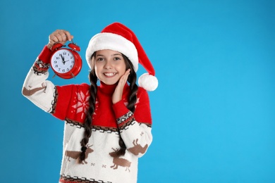 Girl in Santa hat with alarm clock on light blue background, space for text. New Year countdown