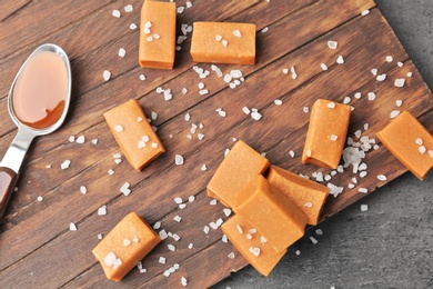 Photo of Delicious caramel sauce in spoon and  candies with salt on wooden board