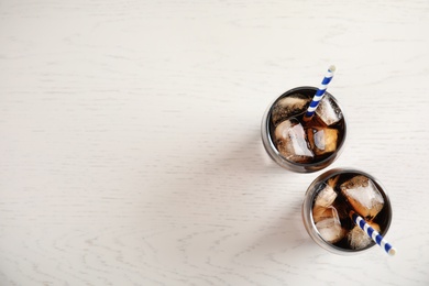 Glasses of refreshing cola with ice cubes and straws on white wooden background, top view. Space for text