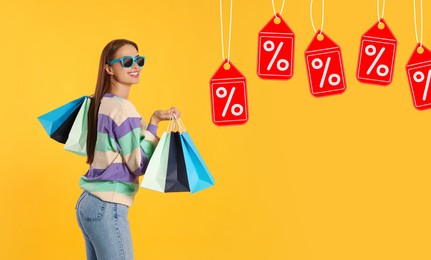 Image of Discount offer. Happy woman with paper shopping bags looking at labels with percent signs on orange background