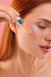 Beautiful young woman applying cosmetic serum onto her face on pink background, closeup