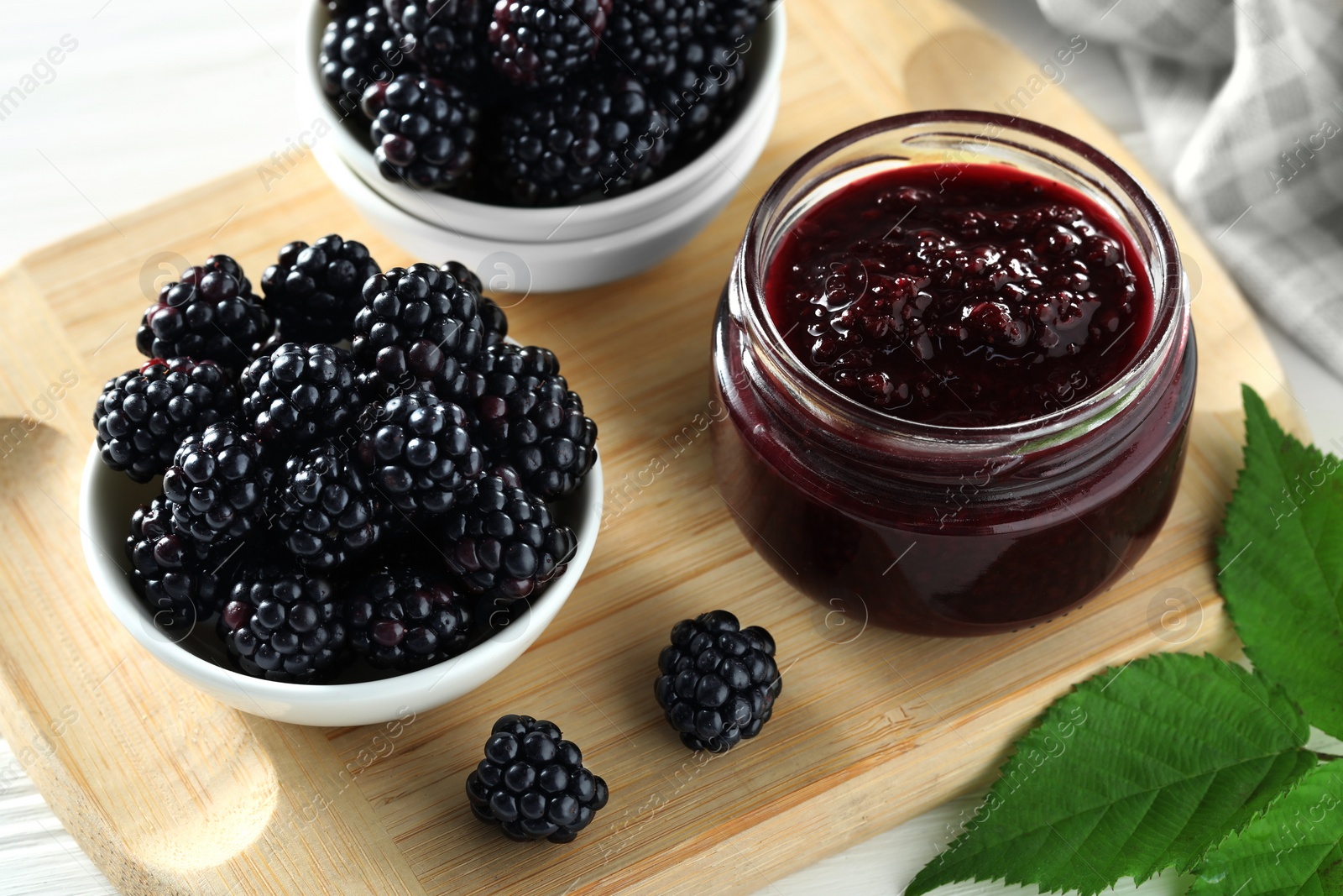 Photo of Fresh ripe blackberries, tasty jam and leaves on wooden board, closeup
