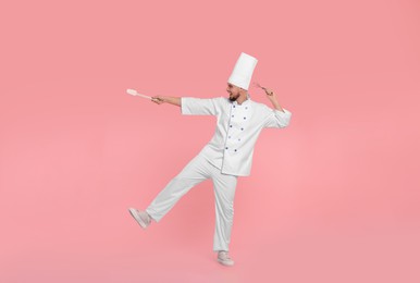 Photo of Happy professional confectioner in uniform having fun with spatula and whisk on pink background