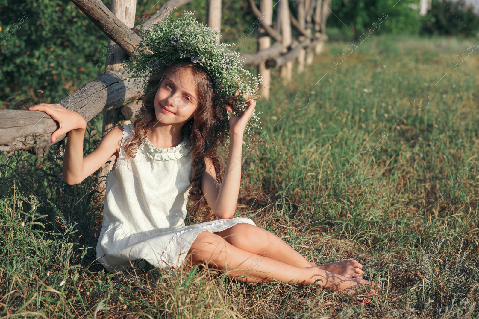 Photo of Cute little girl wearing wreath made of beautiful flowers near wooden fence outdoors on sunny day