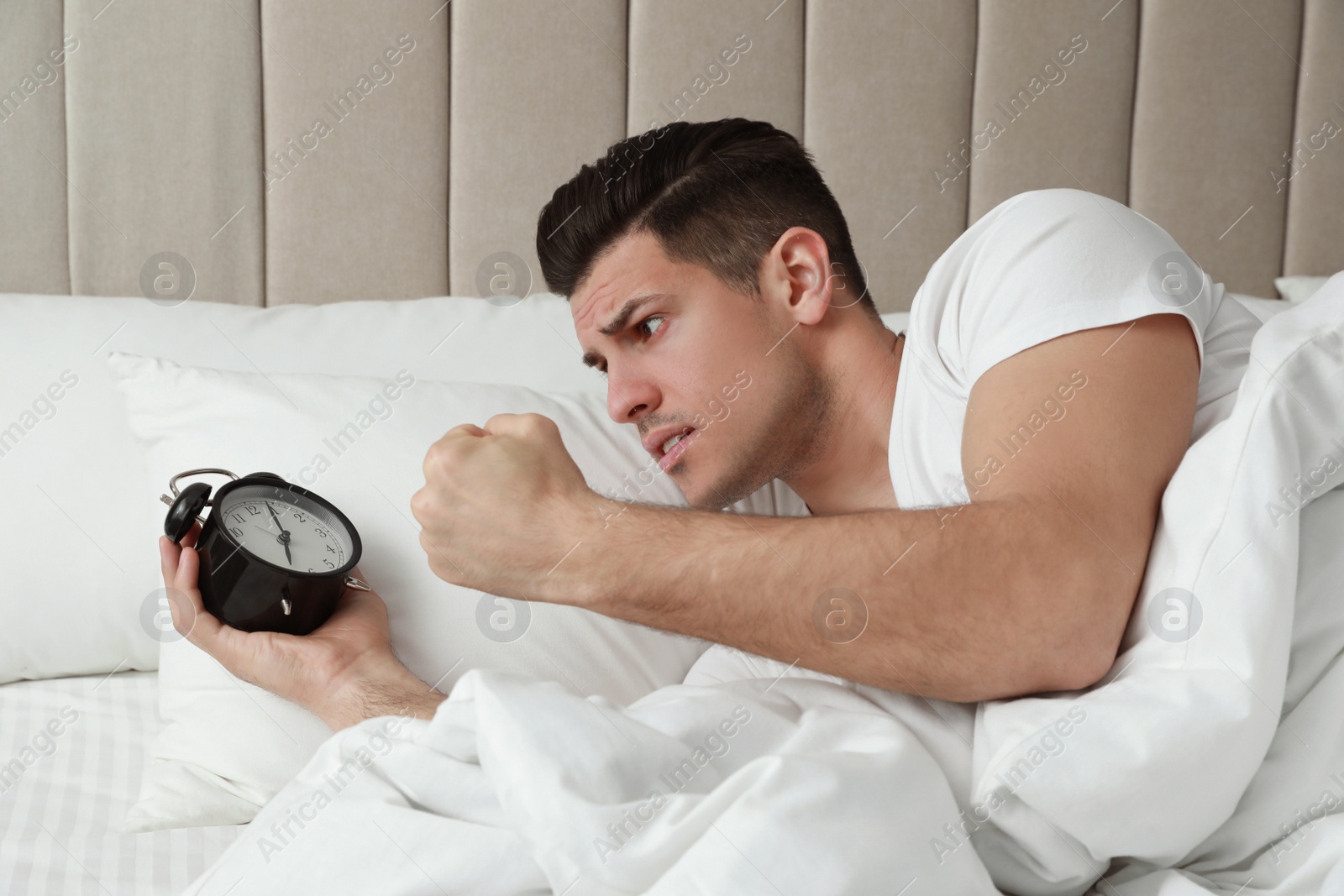 Photo of Emotional man with alarm clock in bed. Being late because of oversleeping