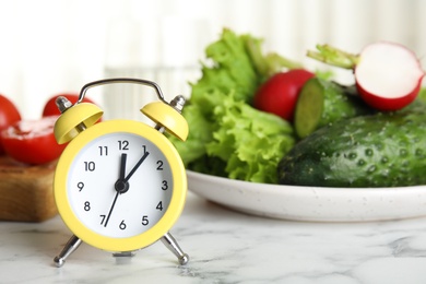 Photo of Alarm clock and vegetables on white marble table. Meal timing concept