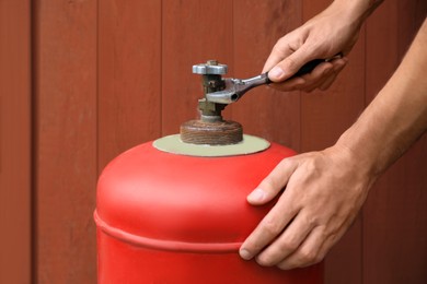 Photo of Man with adjustable wrench opening red gas cylinder near brown wooden wall, closeup