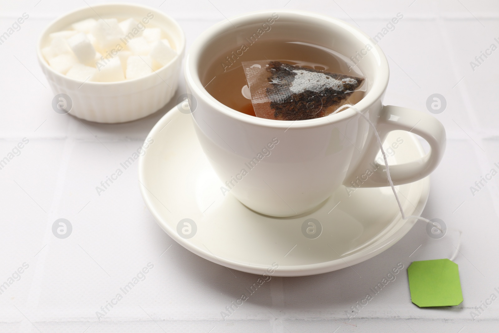 Photo of Tea bag in cup with hot drink and bowl with sugar on white tiled table, closeup
