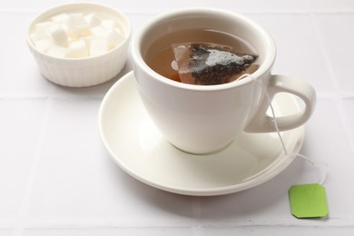 Tea bag in cup with hot drink and bowl with sugar on white tiled table, closeup