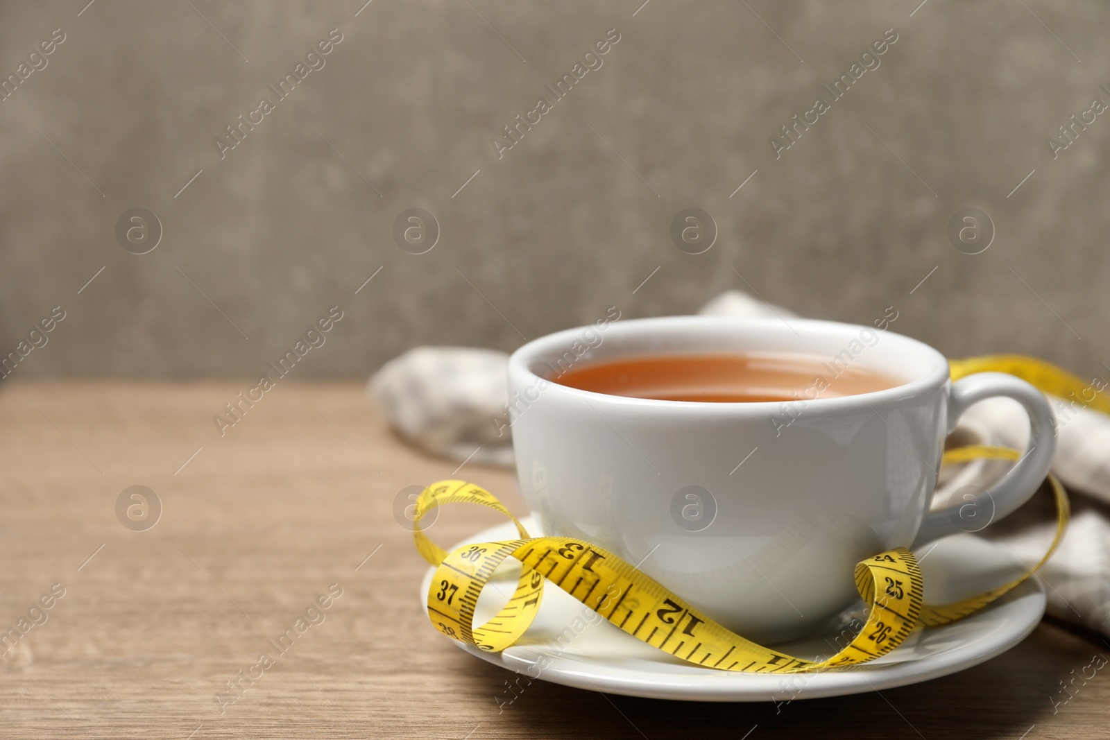 Photo of Cup of herbal diet tea and measuring tape on wooden table against grey wall, space for text. Weight loss concept