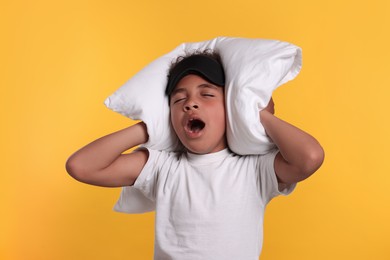 Photo of Boy with sleep mask covering his ears with pillow and yawning on yellow background. Insomnia problem