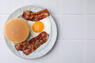 Photo of Plate with tasty pancakes, fried egg and bacon on white tiled table, top view. Space for text