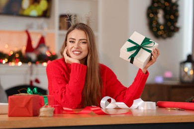 Photo of Beautiful young woman in deer headband with Christmas gift at table in room