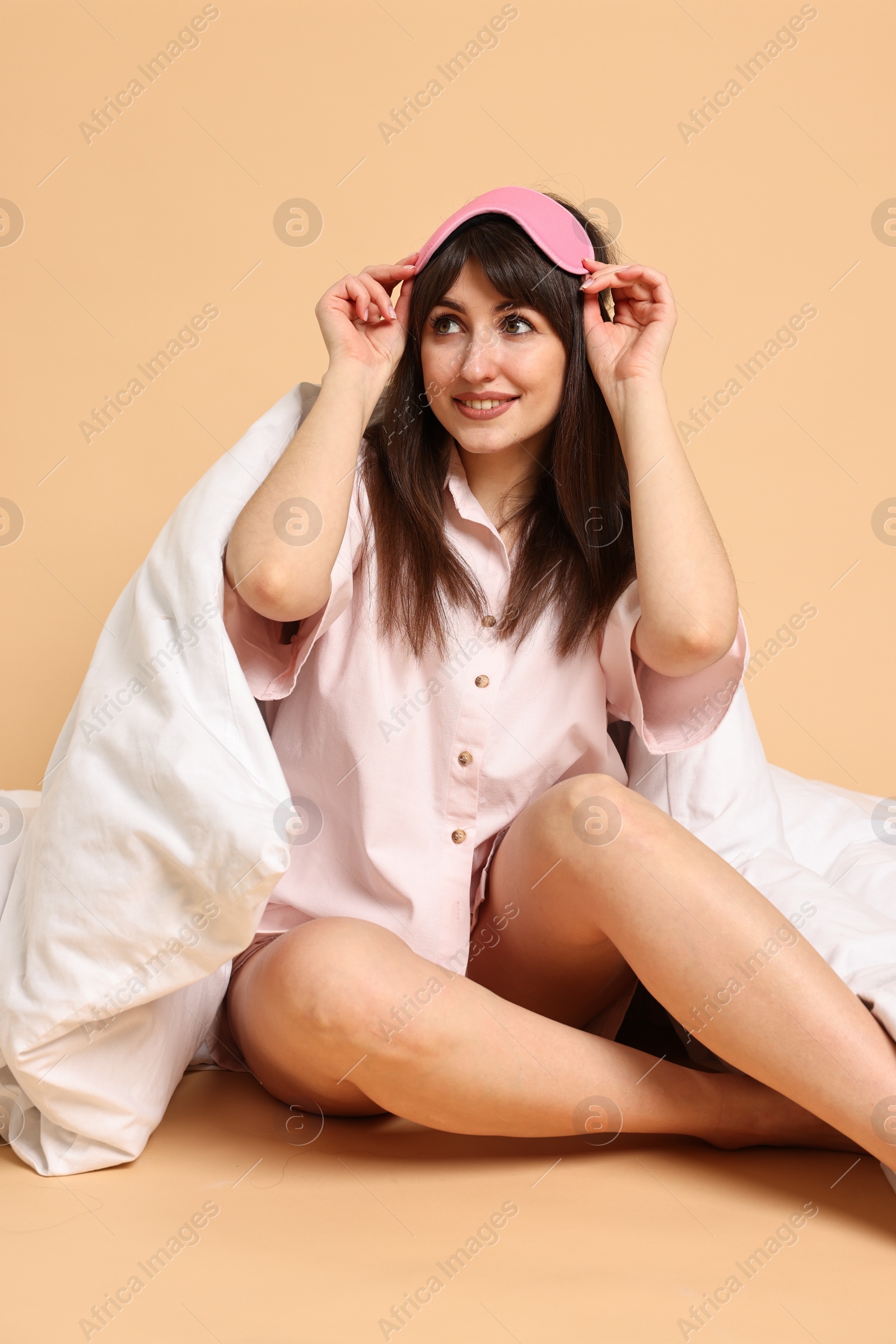 Photo of Happy woman with pyjama and sleep mask wrapped in blanket on beige background