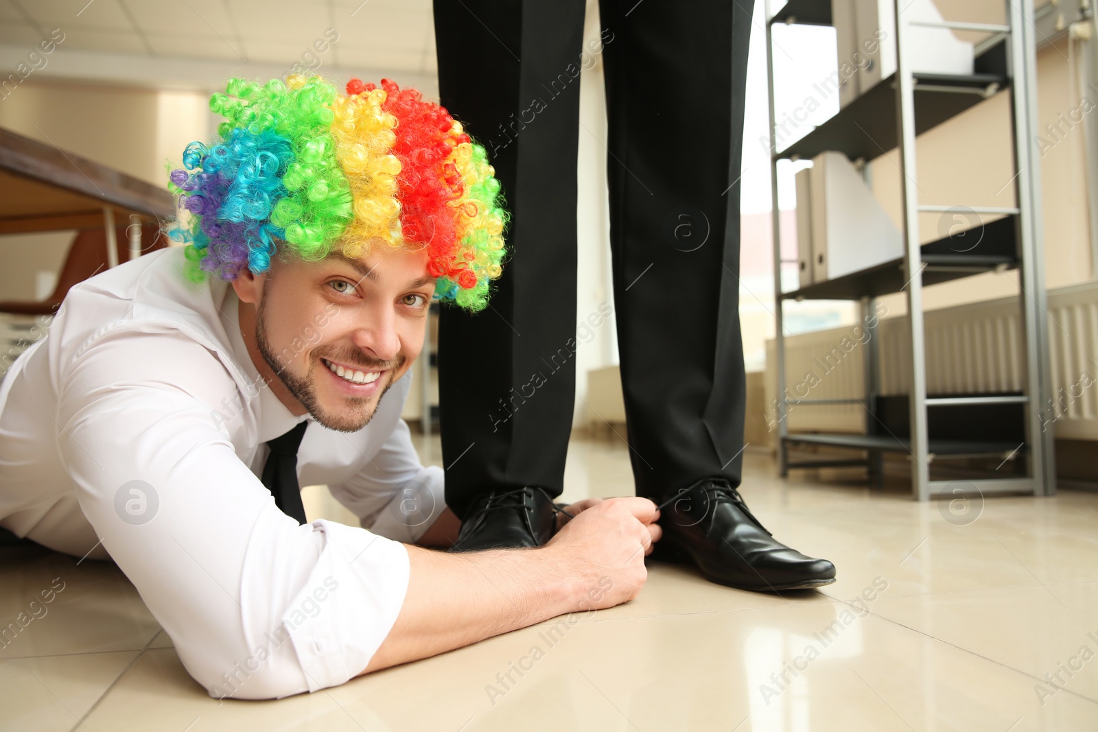 Photo of Man with clown wig tying shoe laces of his colleague together in office, closeup. Funny joke