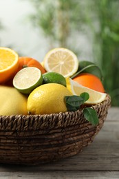 Photo of Different fresh citrus fruits and leaves in wicker basket on wooden table against blurred background, closeup