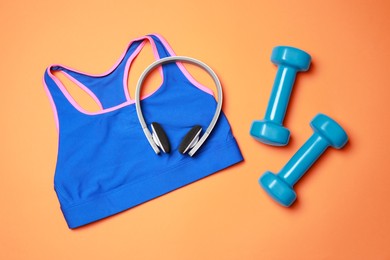 Photo of Stylish sports bra, dumbbells and headphones on orange background, flat lay