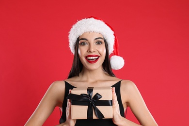 Photo of Excited woman in black dress and Santa hat holding Christmas gift on red background