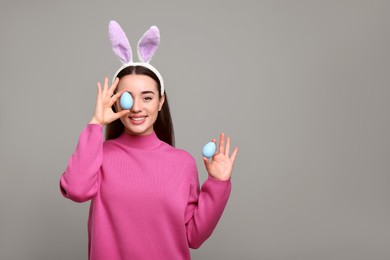 Photo of Happy woman in bunny ears headband holding painted Easter eggs on grey background. Space for text.