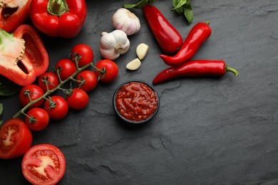 Delicious adjika sauce in bowl and ingredients on black table, flat lay