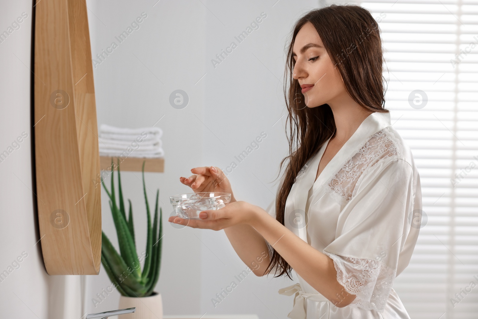 Photo of Young woman holding bowl of aloe hair mask near mirror in bathroom